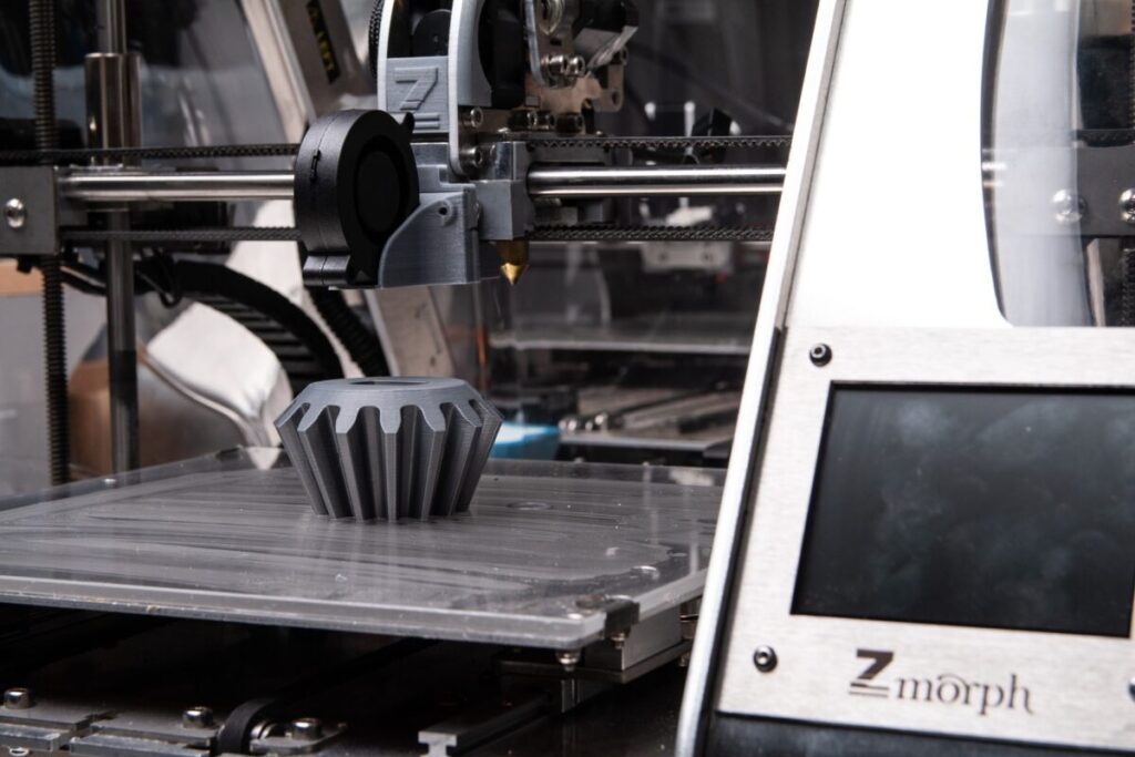 An FDM 3D printed gear is shown sitting on the build plate of an opened 3D printing machine.