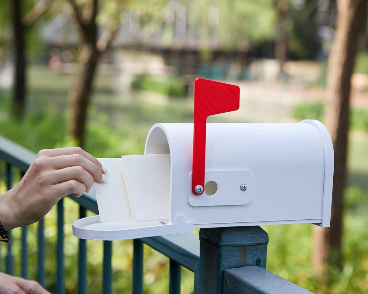 A white mailbox with a red carrier flag in the vertical position, is shown sitting atop a dark colored fence outdoors. The mailbox is entirely 3D printed out of ASA material.