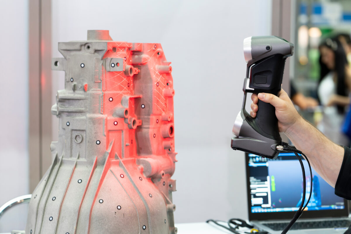 A light-colored bell housing for an automotive transmission is scanned with a red laser-light hand-held 3D scanner. A laptop in the background shows the 3D model of the scan being generated in real time.